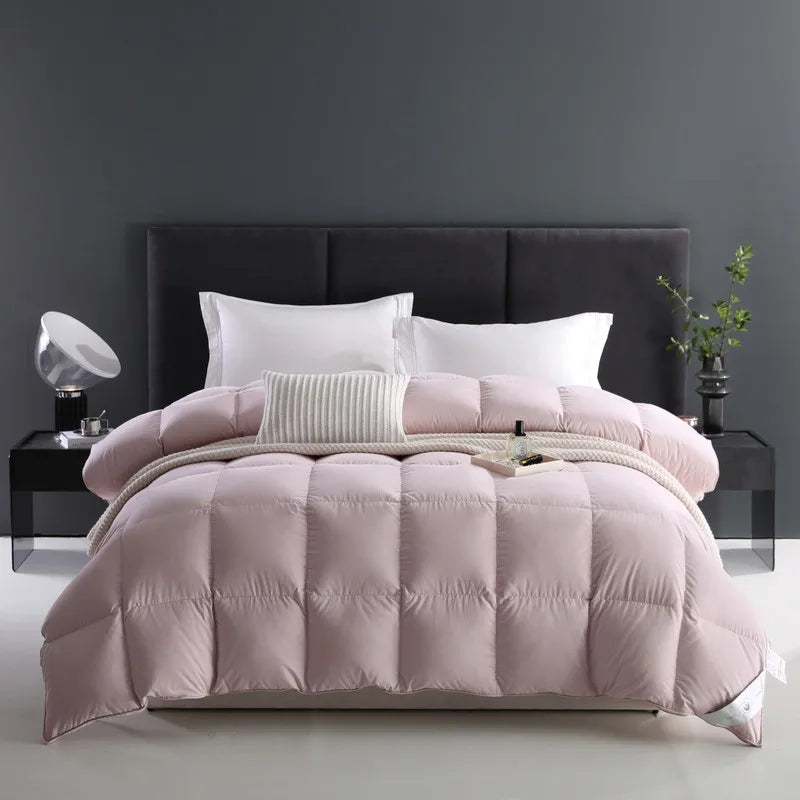 A neatly made bed featuring a 100% cotton comforter in light pink with premium white goose down fill, offering a breathable and fluffy texture. The solid color design complements the arranged pillows, creating an inviting and cozy look. Available in Queen and King sizes.